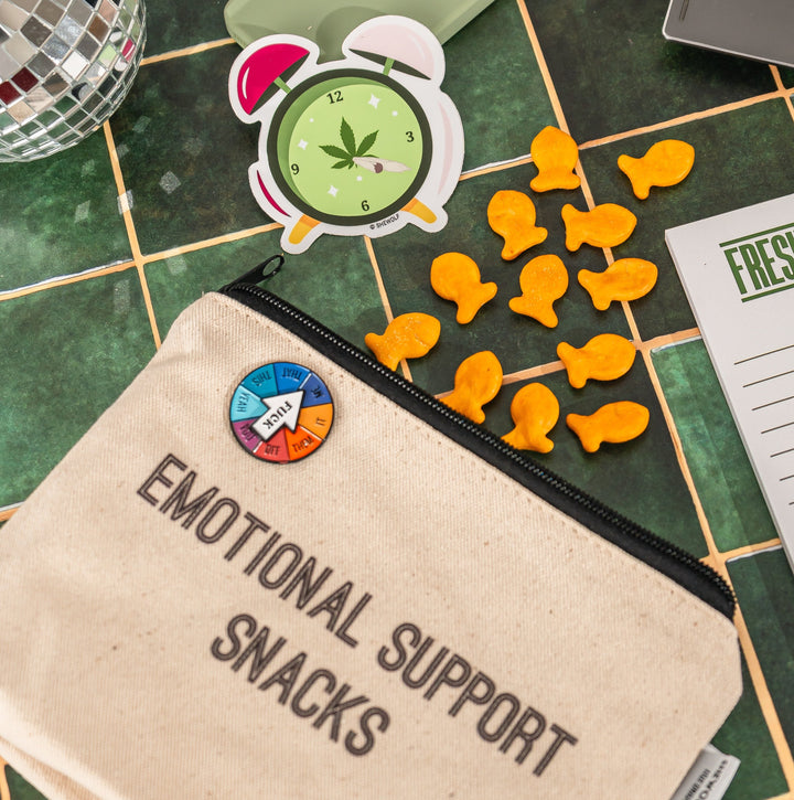group picture of products, including an interactive spinner that says "fuck" with different options and a zippered canvas pouch with "emotional support snacks" written on it