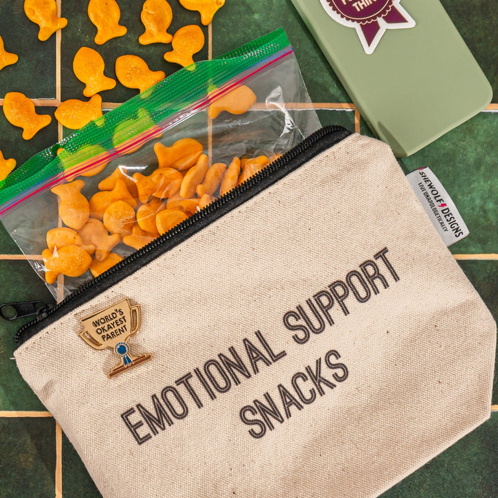 Canvas pouch that says "emotional support snacks" and a gold enamel pin in the shape of a trophy that says "world's okayest parent"